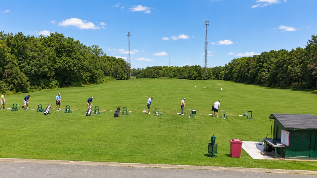 Golfers practicing their swings at individual stations on the driving range at Belmont Lake Golf Club, with tree-lined fairway and small shelter building