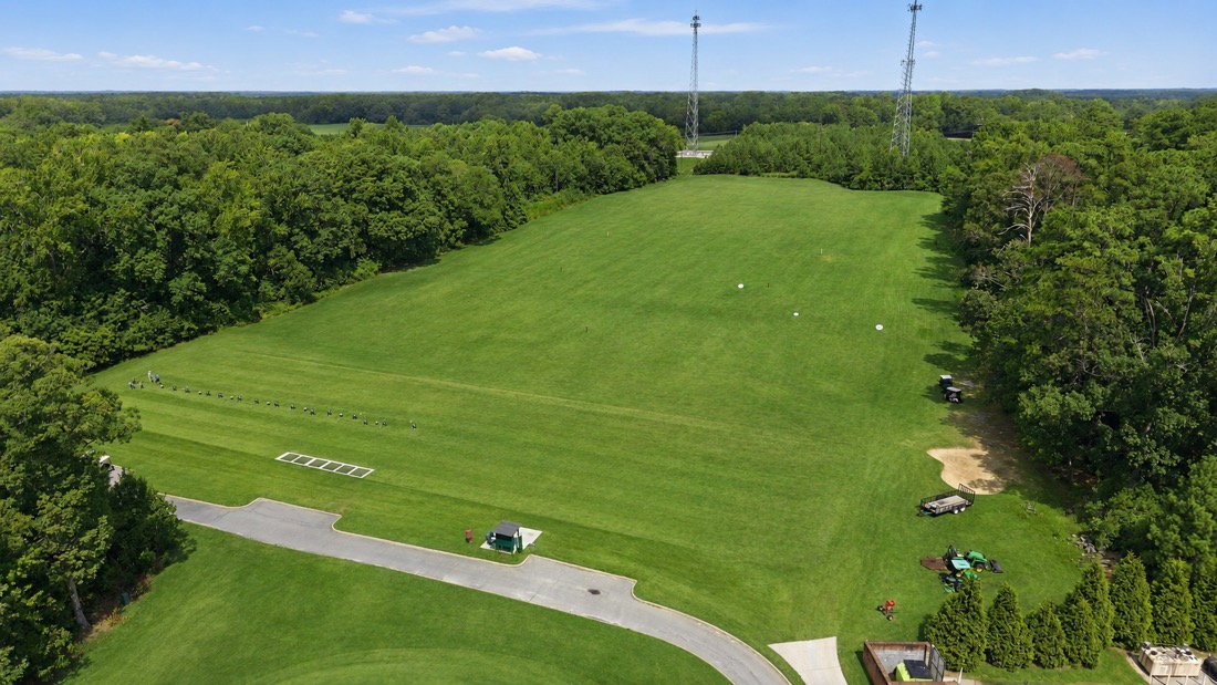 Aerial view of the driving range at Belmont Lake Golf Club, showing a wide grass tee line and landing area surrounded by trees
